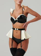 Black and white latex rubber lingerie set including a bra and suspender belt with frills and bows, and matching hipster briefs. Worn by a model also wearing black stockings, on a light grey background.