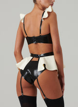Back view of a black and white latex rubber lingerie set including a bra and suspender belt with frills and bows, and matching hipster briefs. Worn by a model also wearing black stockings, on a light grey background.