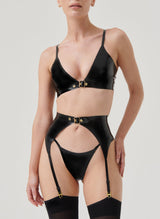 A model wearing a black latex lingerie set with gold buckles, including a bra, suspender belt and G-string, all by William Wilde. Model also wears black nylon stockings.
