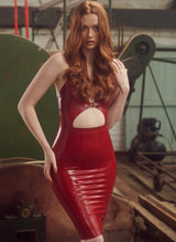 A model with ginger hair wearing a plum coloured latex pencil dress with a front cutout and gold buckle detail, with adjustable shoulder straps.