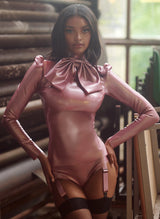 A model wearing a pearlsheen pink latex blouse with XL pussy bow and matching suspender knickers. Black fishnet stockings complete the look.