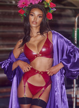 A model wearing a plum latex lingerie set with gold buckles, including a bra, suspender belt and G-string, all by William Wilde. Model also wears a floral headband, purple satin robe and fishnet stockings.