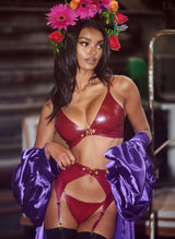 A model wearing a plum latex lingerie set with gold buckles, including a bra, suspender belt and G-string, all by William Wilde. Model also wears a floral headband, purple satin robe and stockings.