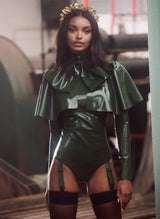 A model wearing an olive green latex cape with matching pussy bow, blouse and suspender knickers. Also worn is a gold headband and fishnet stockings.