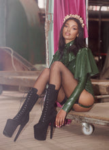 A model sitting in a warehouse like setting, wearing an olive green latex cape, blouse and knickers, with a gold headband and black high heeled boots.