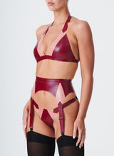 Side view of a model wearing a plum latex lingerie set with Semi-Transparent pink inserts. The latex lingerie set includes a bikini-style halter bra, four strap suspender belt and thong. Model also wears black nylon stockings.