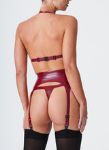 Back view of a model wearing a plum latex lingerie set. The latex lingerie set includes a bikini-style halter bra, four strap suspender belt and thong. Model also wears black nylon stockings.