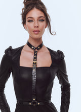 A model wearing a soft black leatherette pencil dress with square neckline and gathered sleeve heads. Also wearing a black leather harness with gold buckles.