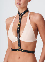 A model wearing a peach latex bra and the Natalia Harness, which is made from shiny black leather with gold metal buckles, rings and eyelets.
