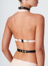Back view of a model wearing peach latex bra and black leather harness with gold fastenings.
