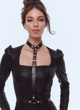A model wearing a black leatherette dress with a black leather harness over the top. Fastenings are of gold metal hardware.