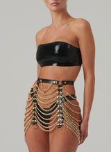 A model wearing a black latex bandeau top and matching latex high leg thong. Worn over the top is a harness skirt made from black patent leather and gold chain, with gold hardware. All items designed by William Wilde.