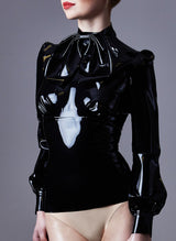 A model with red lipstick on a grey background wearing a high-necked, long-sleeved black latex blouse, with bishop sleeves and pleated details. A removable XL pussy bow is worn at the neck.