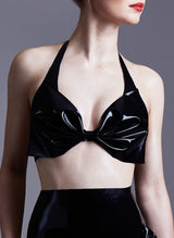A model with red lipstick wears a glossy black latex halter neck bra with a large bow at the front cups. Matching high waisted knickers also shown.