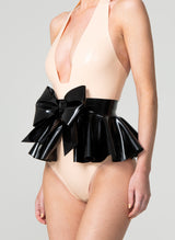 A person wearing a peach latex bodysuit and a black latex rubber belt with a large bow and flared peplum, fastened with press studs at the back.