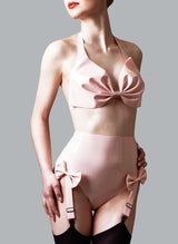 A model wearing a rose-petal pink latex lingerie set with handcrafted bows, comprising of a halter bra and high-waisted suspender knickers. Worn here with black stockings.