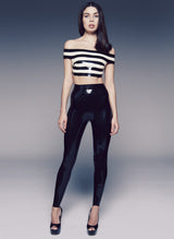 A model on a grey background wears black latex high waisted leggings, with a black and white striped off-the-shoulder cropped top.