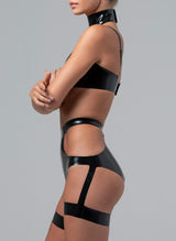 Side shot of a model wearing a black latex bra and briefs. The bra has skinny shoulder straps and a collar attached, the briefs have cut-outs at the hips and thigh bands attached with straps. By designer William Wilde.