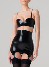 A model wearing a shiny black latex rubber bra and matching girdle with suspender clips at hem. Both by designer William Wilde. Worn here with black nylon stockings.