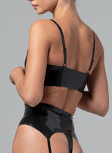 back of a black latex bra top with chunky silver metal zip at the front and adjustable shoulder straps. The garment has a glossy finish and is modelled by a woman. Matching suspender belt also visible.