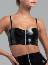 A black latex bra top with chunky silver metal zip at the front and adjustable shoulder straps. The garment has a glossy finish and is modelled by a woman. Matching suspender belt also visible.