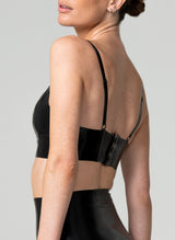 Back shot of a model wearing a black latex bra with a V neckline and adjustable shoulder straps. Fastening is with press studs. Matching latex suspender belt also in shot, all by designer William Wilde.