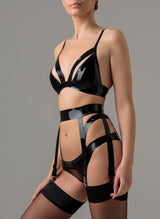 A model on a grey background wears a glossy black latex lingerie set, including bra and six strap suspender belt, both with cut out details and adjustable straps. Latex hipster briefs also included. Shown with black stockings, all by designer William Wilde.
