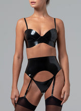 A black latex rubber lingerie set worn by a model with matching stockings on a grey background. The latex lingerie set includes a bra and wide suspender belt, with matching G-string, all with skinny straps.