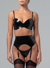 A black latex rubber lingerie set worn by a model with matching stockings on a grey background. The latex lingerie set includes a bra and wide suspender belt, with matching G-string, all with skinny straps.
