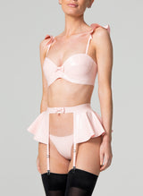 A model wearing a baby pink latex lingerie set, including bra with frilled shoulders, suspender belt with peplum and g-string, all by designer William Wilde. The model also wears black nylon stockings.