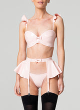 A model wearing a baby pink latex lingerie set, including bra with frilled shoulders, suspender belt with peplum and g-string, all by designer William Wilde. The model also wears black nylon stockings.
