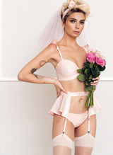 A model with tattoos wearing a baby pink latex lingerie set, including bra with frilled shoulders, suspender belt with peplum and latex briefs, all by designer William Wilde. The model also wears black nylon stockings, and a pink latex hair bow.