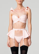 A model wearing a baby pink latex lingerie set, including bra with frilled shoulders, suspender belt with peplum and g-string, all by designer William Wilde. The model also wears black nylon stockings.