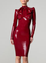 A model wearing a skintight, plum latex knee-length dress with long sleeves, high collar and an XL pussy bow at the neck. The dress features pleated sleeve heads and undersbust seam with bust-gathers.