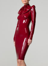 A model wearing a skintight, plum latex knee-length dress with long sleeves, high collar and an XL pussy bow at the neck. The dress features pleated sleeve heads and undersbust seam with bust-gathers.