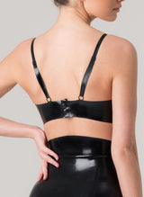 Back of a model wearing a black latex bra with shoulder straps. The bra is featured in a close-up shot, showcasing its design and fit. Matching latex skirt also worn, by designer William Wilde.