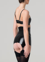 Back view of a model wearing a shiny black latex rubber bra and matching girdle with suspender clips at hem. Both by designer William Wilde. Worn here with black nylon stockings.