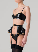 Side view of a model wearing a black latex rubber lingerie set with gold latex trim. Set includes a bra with gold trim, featuring adjustable shoulder straps, and a mini bow at the front. Matching latex suspender belt with peplum and hipster briefs also in the set, all by designer William Wilde.