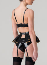 Back view of a model wearing a black latex rubber lingerie set with gold latex trim. Set includes a bra with gold trim, featuring adjustable shoulder straps, and a mini bow at the front. Matching latex suspender belt with peplum and hipster briefs also in the set, all by designer William Wilde.