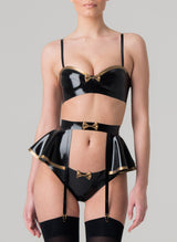 A model wearing a black latex rubber lingerie set with gold latex trim. Set includes a bra with gold trim, featuring adjustable shoulder straps, and a mini bow at the front. Matching latex suspender belt with peplum and hipster briefs also in the set, all by designer William Wilde.