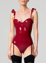 A person wearing a shiny plum latex bodysuit with frilled shoulder detail and mini bow at the front, paired with black opaque stockings.