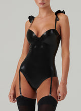 A person wearing a shiny black latex bodysuit with frilled shoulder detail and mini bow at the front, paired with black opaque stockings.