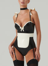 A model wearing a black and white latex costume with frilled shoulders, a mini bow at the front, adjustable straps, an apron, a choker, and a hair bow. Worn here with black stockings.