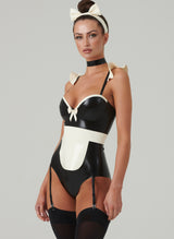 A model wearing a black and white latex costume with frilled shoulders, a mini bow at the front, adjustable straps, an apron, a choker, and a hair bow. Worn here with black stockings.