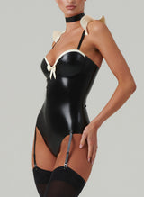 A model wearing a black and white latex costume with frilled shoulders, a mini bow at the front, adjustable straps, a choker, and a hair bow. Worn here with black stockings.