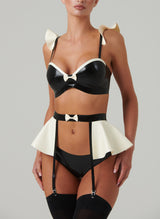 Black and white latex rubber lingerie set including a bra and suspender belt with frills and bows, and matching hipster briefs. Worn by a model also wearing black stockings, on a light grey background.