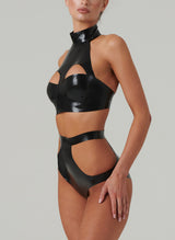 A black latex bra top by William Wilde with a high collar and keyhole cut-outs, worn by a model wearing black latex knickers with hip cut-outs, against a neutral background.