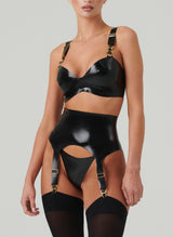 A black shiny latex lingerie set worn by a model. The set includes a bra and suspender belt, both with gold metal sliders, press studs and suspender clips. Worn with matching G-String and black opaque stockings. All by designer William Wilde