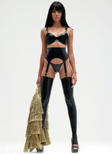 A black shiny latex lingerie set worn by a model. The set includes a bra and suspender belt, both with gold metal sliders, press studs and suspender clips. Worn with matching G-String and black latex stockings. All by designer William Wilde. The model holds a gold tinsel jacket.