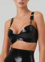 A black latex bra with gold suspender clasps and rings, featuring adjustable shoulder straps and fastened at the back. The model is wearing the bra with a matching latex lingerie set.